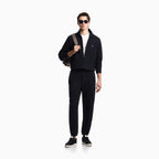 A man modeling a black zip-up jacket and matching black track pants, styled with a white t-shirt, black sneakers, sunglasses, and a gray shoulder bag against a plain white background. Emporio Armani Sweatshirt available at Sakks Menswear in Southampton.