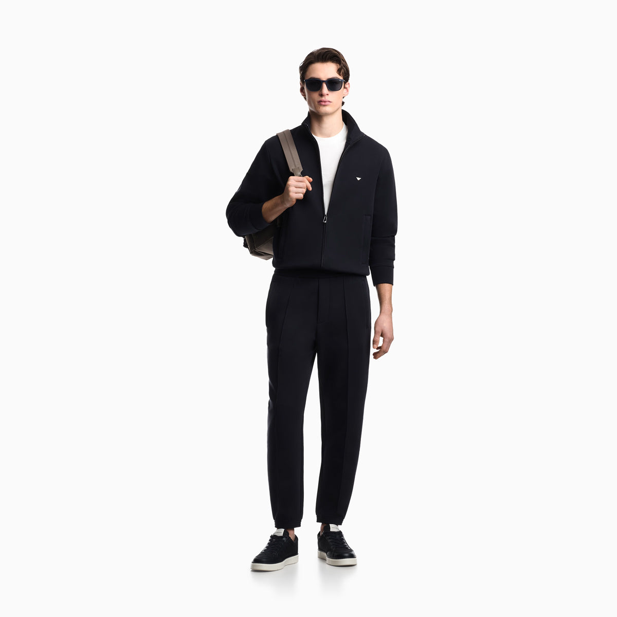 A man modeling a black zip-up jacket and matching black track pants, styled with a white t-shirt, black sneakers, sunglasses, and a gray shoulder bag against a plain white background. Emporio Armani Sweatshirt available at Sakks Menswear in Southampton.