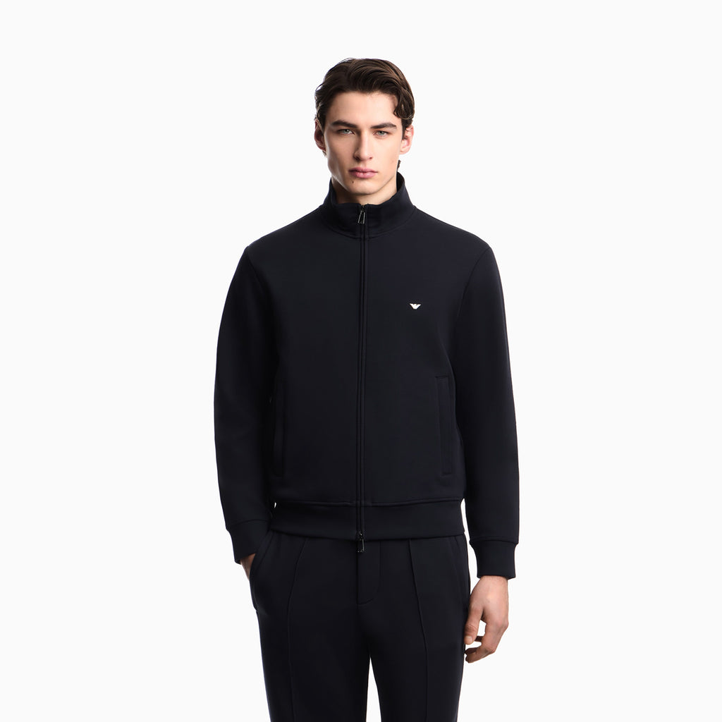 A man modeling a black zip-up track jacket with a small white embroidered logo on the left chest, paired with matching black track pants, against a plain white studio background. Emporio Armani Sweatshirt available at Sakks Menswear in Southampton.