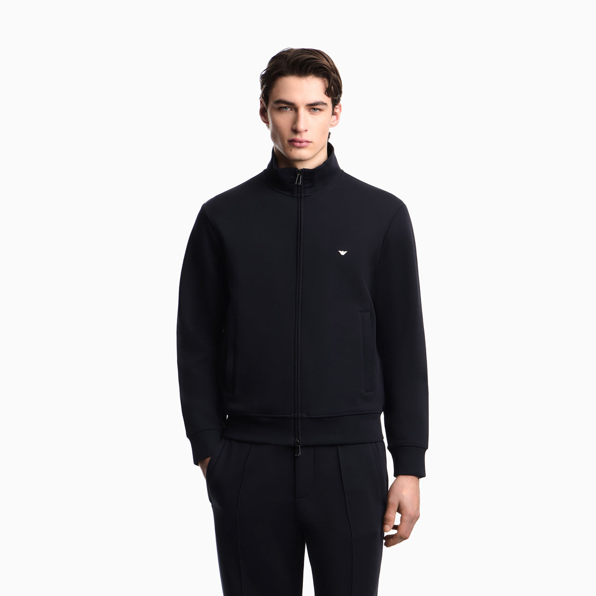 A man modeling a black zip-up track jacket with a small white embroidered logo on the left chest, paired with matching black track pants, against a plain white studio background. Emporio Armani Sweatshirt available at Sakks Menswear in Southampton.