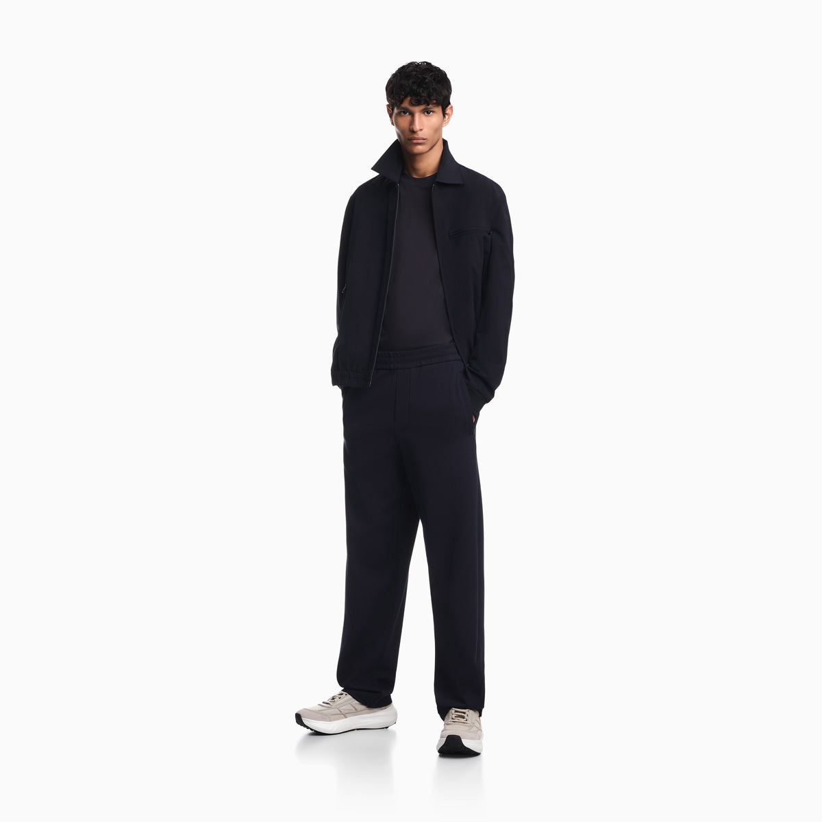 A male model stands in a studio wearing a dark navy blue jacket, matching sweatshirt, and trousers, paired with light gray sneakers, against a solid white background. Emporio Armani Jacket available at Sakks Menswear in Southampton.