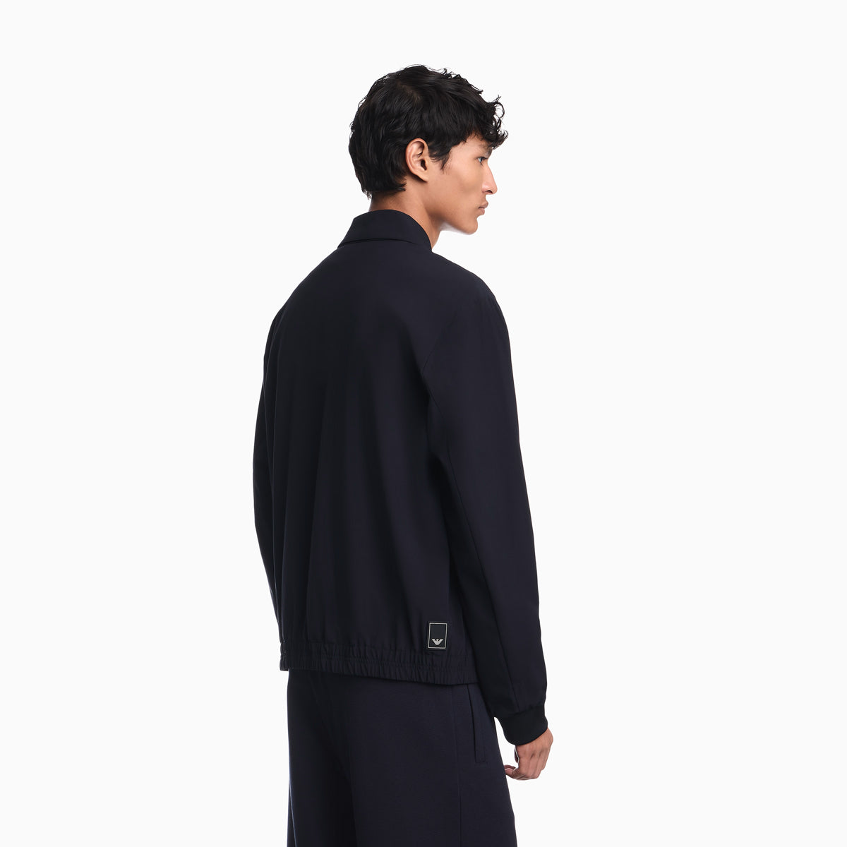 A man shown from the back wearing a dark navy knit zip-up jacket and matching dark trousers against a clean white studio background. Emporio Armani Jacket available at Sakks Menswear in Southampton.