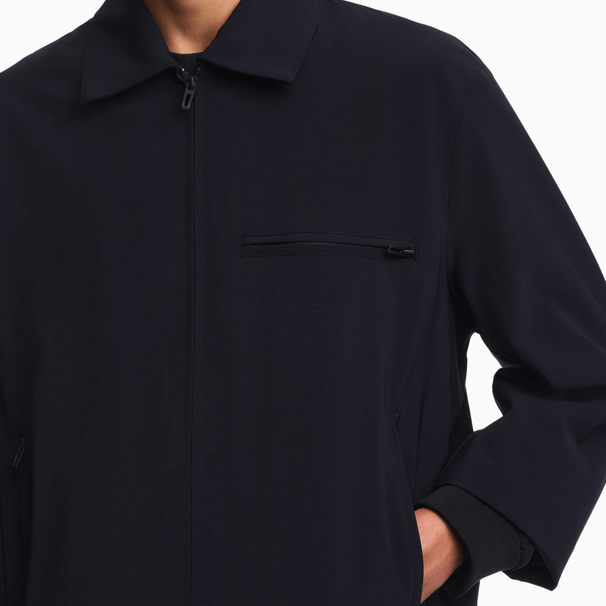 A close-up of a person wearing a dark navy zip-front jacket with a collar and a zippered chest pocket, shown against a plain white background. Emporio Armani Jacket available at Sakks Menswear in Southampton.
