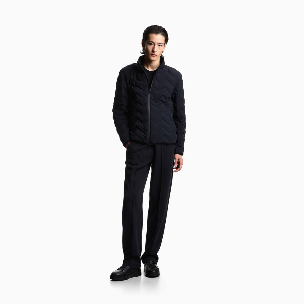 A model stands against a plain white studio background wearing a dark navy quilted zip-up jacket, matching dark trousers, and black shoes. Emporio Armani Down Jacket available at Sakks Menswear in Southampton.
