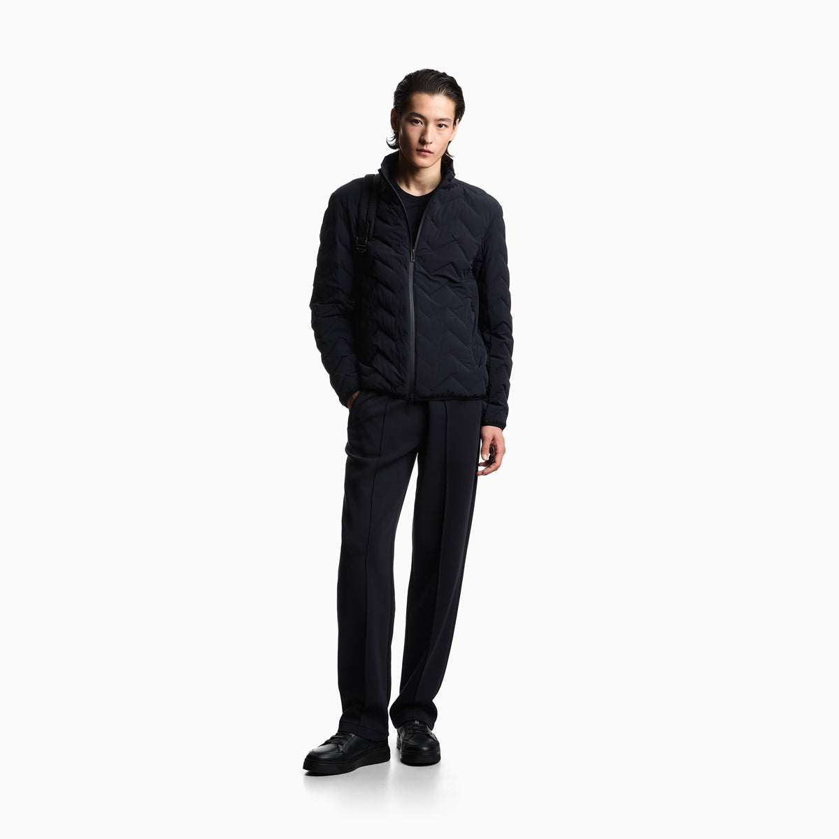 A model stands against a plain white studio background wearing a dark navy quilted zip-up jacket, matching dark trousers, and black shoes. Emporio Armani Down Jacket available at Sakks Menswear in Southampton.