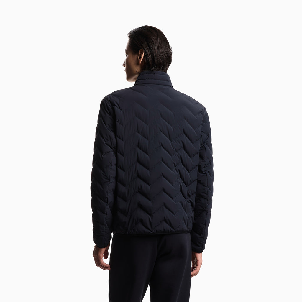 A man viewed from the back wearing a dark navy quilted puffer jacket with a distinctive chevron pattern and black pants, set against a plain white studio background. Emporio Armani Down Jacket available at Sakks Menswear in Southampton.
