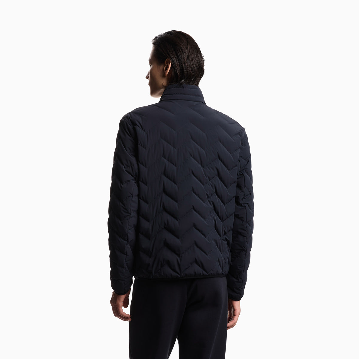 A man viewed from the back wearing a dark navy quilted puffer jacket with a distinctive chevron pattern and black pants, set against a plain white studio background. Emporio Armani Down Jacket available at Sakks Menswear in Southampton.