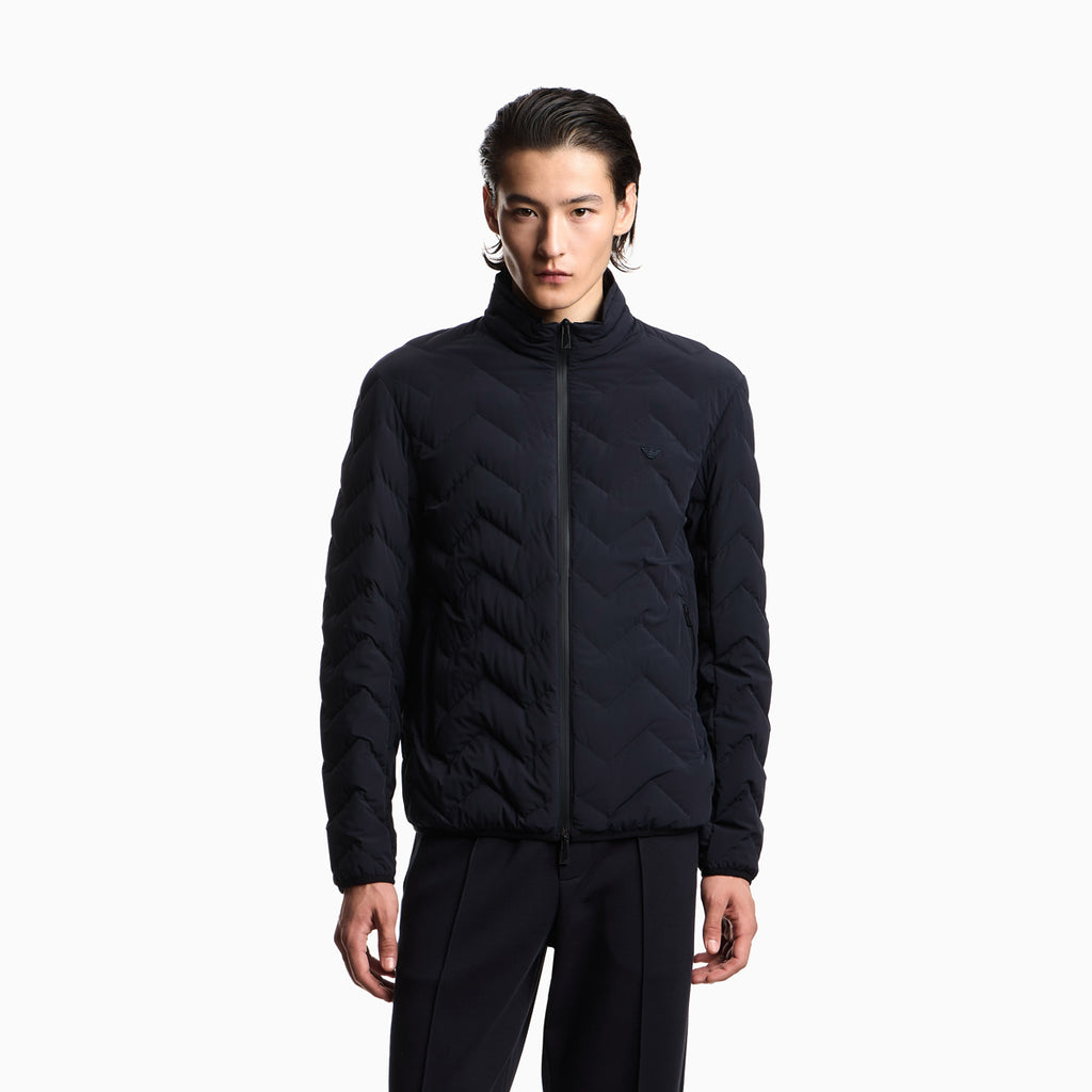 A man wearing a black quilted zip-up jacket with a stand-up collar and black pants, standing against a plain white studio background. Emporio Armani Down Jacket available at Sakks Menswear in Southampton.