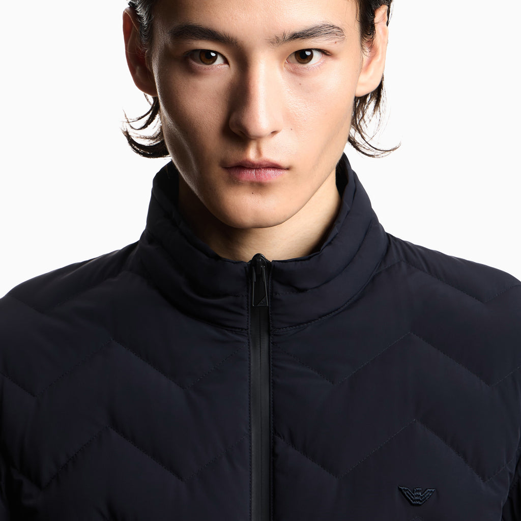 Close-up of a model wearing a dark navy blue quilted zip-up jacket with a high collar and a small embroidered logo on the chest, set against a plain white background. Emporio Armani Down Jacket available at Sakks Menswear in Southampton.
