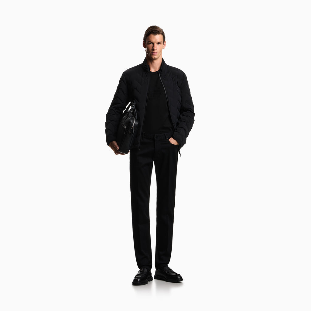 A male model stands against a plain white studio background wearing a black bomber jacket over a black shirt and black trousers, holding a black leather bag. Emporio Armani Jeans available at Sakks Menswear in Southampton.