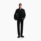 A male model stands against a plain white studio background wearing a black bomber jacket over a black shirt and black trousers, holding a black leather bag. Emporio Armani Jeans available at Sakks Menswear in Southampton.