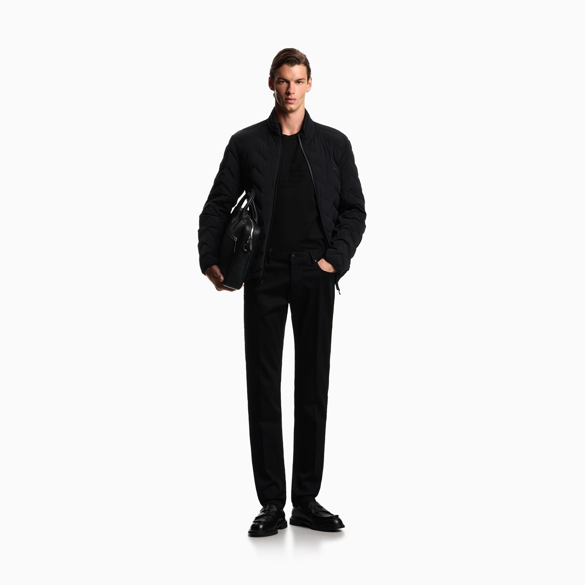 A male model stands against a plain white studio background wearing a black bomber jacket over a black shirt and black trousers, holding a black leather bag. Emporio Armani Jeans available at Sakks Menswear in Southampton.