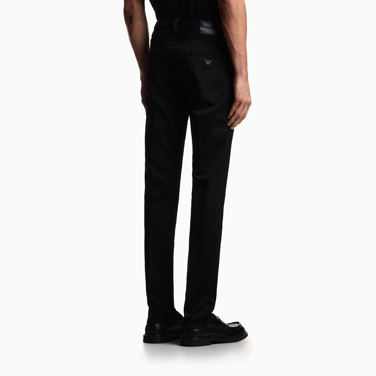 A model shown from the back wearing black slim-fit trousers, a black shirt, and black loafers against a plain white studio background. Emporio Armani Jeans available at Sakks Menswear in Southampton.