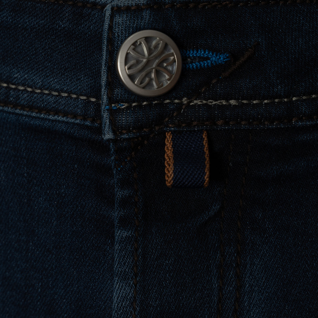 Close-up detail of dark indigo denim jeans showing the button closure, contrast stitching, and a small blue and brown brand tab. Jacob Cohen Jeans available at Sakks Menswear in Southampton.