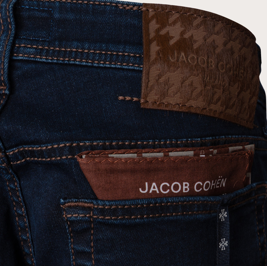 Close-up of the back waistband and pocket area of dark blue denim jeans, showing brown leather brand patches with JACOB COHEN text. Jacob Cohen Jeans available at Sakks Menswear in Southampton.