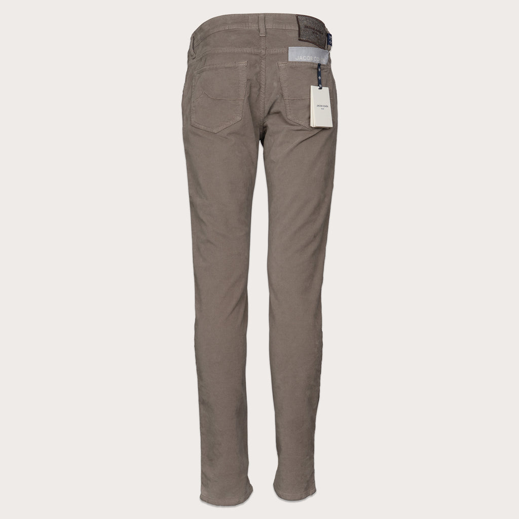 A pair of slim-fit trousers in a muted taupe-brown color, shown from the back on a light beige background. The trousers feature a five-pocket design, a visible waistband, and a white product tag hanging from the right pocket. Jacob Cohen Trousers available at Sakks Menswear in Southampton.