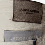 Close-up of a light beige woven fabric garment showing two Jacob Cohen labels: a dark brown textured back label reading 'JACOB COHÉN MILANO' and a lighter gray front pocket label. Jacob Cohen Jeans available at Sakks Menswear in Southampton.