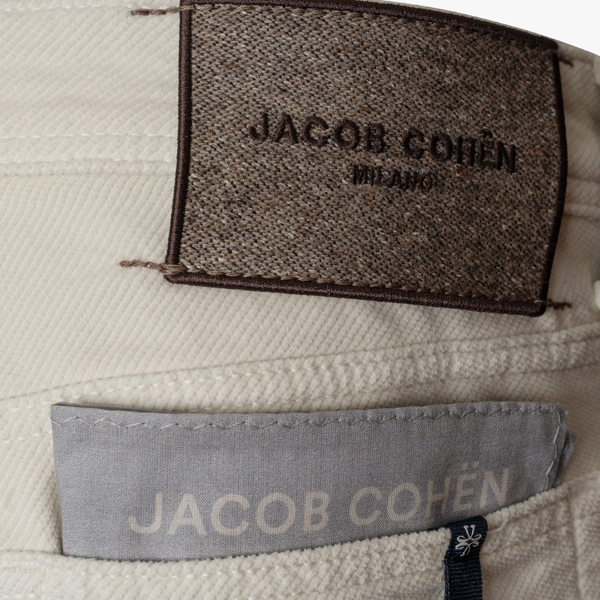 Close-up of a light beige woven fabric garment showing two Jacob Cohen labels: a dark brown textured back label reading 'JACOB COHÉN MILANO' and a lighter gray front pocket label. Jacob Cohen Jeans available at Sakks Menswear in Southampton.