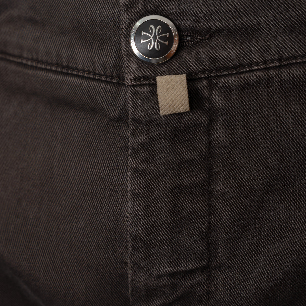 Close-up of dark charcoal gray denim fabric showing a silver metal button with a stylized logo and a small beige fabric brand tab. Jacob Cohen Trousers available at Sakks Menswear in Southampton.