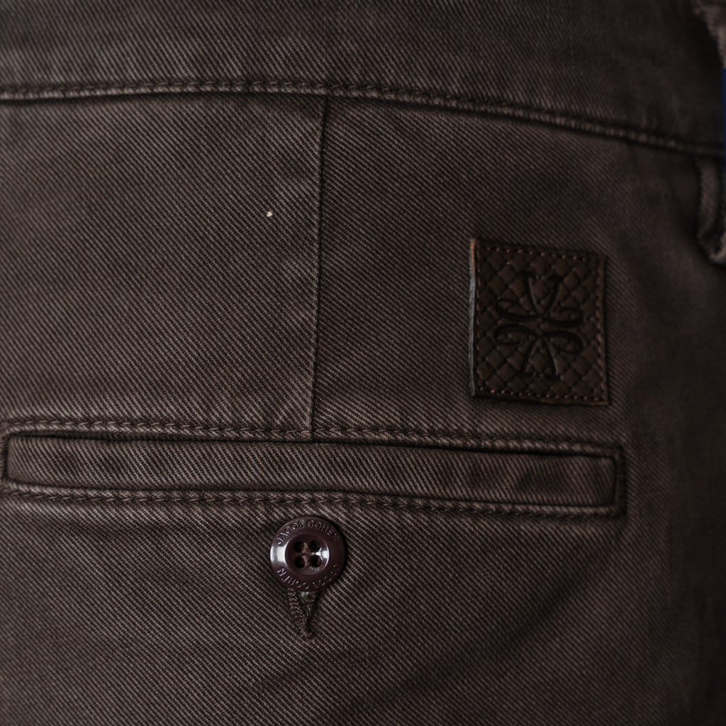 Close-up of the back waistband of dark brown denim jeans, showing a buttoned welt pocket, a small square leather brand patch with an embossed floral design, and the top of a button fly. Jacob Cohen Trousers available at Sakks Menswear in Southampton.