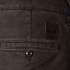 Close-up of the back waistband of dark brown denim jeans, showing a buttoned welt pocket, a small square leather brand patch with an embossed floral design, and the top of a button fly. Jacob Cohen Trousers available at Sakks Menswear in Southampton.