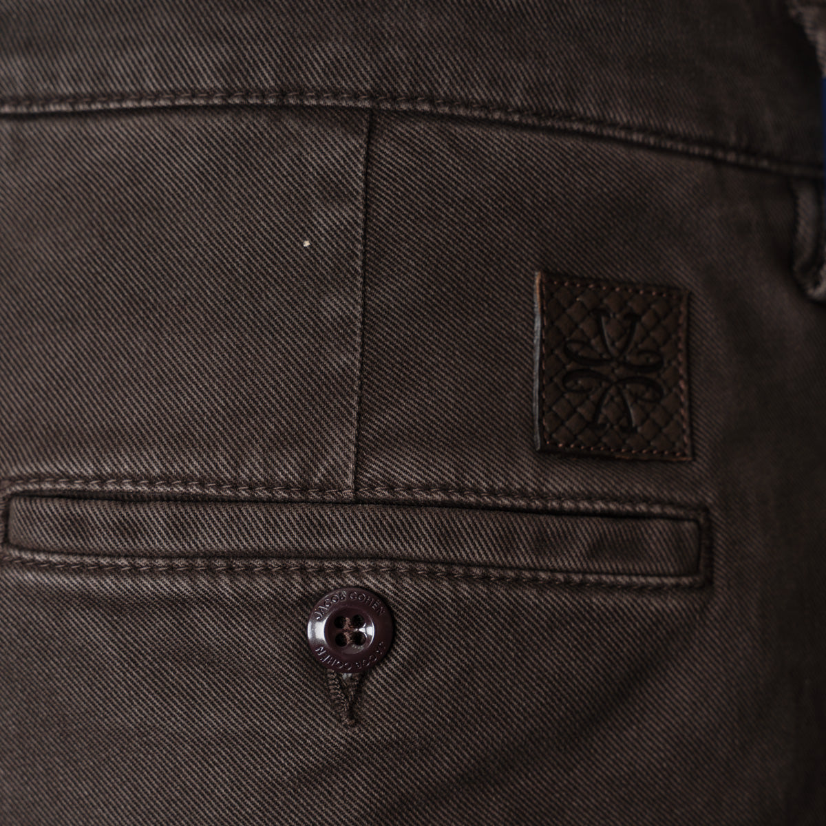 Close-up of the back waistband of dark brown denim jeans, showing a buttoned welt pocket, a small square leather brand patch with an embossed floral design, and the top of a button fly. Jacob Cohen Trousers available at Sakks Menswear in Southampton.