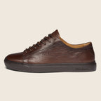 A brown leather low-top sneaker with dark brown laces and a thick dark sole, shown in profile against a light beige background. The brand 'Oliver Sweeney' is visible on the side of the sole. Oliver Sweeney Shoes are available at Sakks Menswear in Southampton.