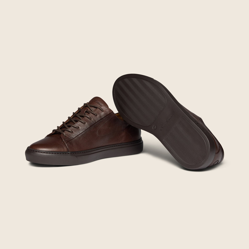 A pair of dark brown leather low-top sneakers, with one shoe standing upright and the other lying on its side to display the sole, set against a solid light beige background. Oliver Sweeney Shoes are available at Sakks Menswear in Southampton.