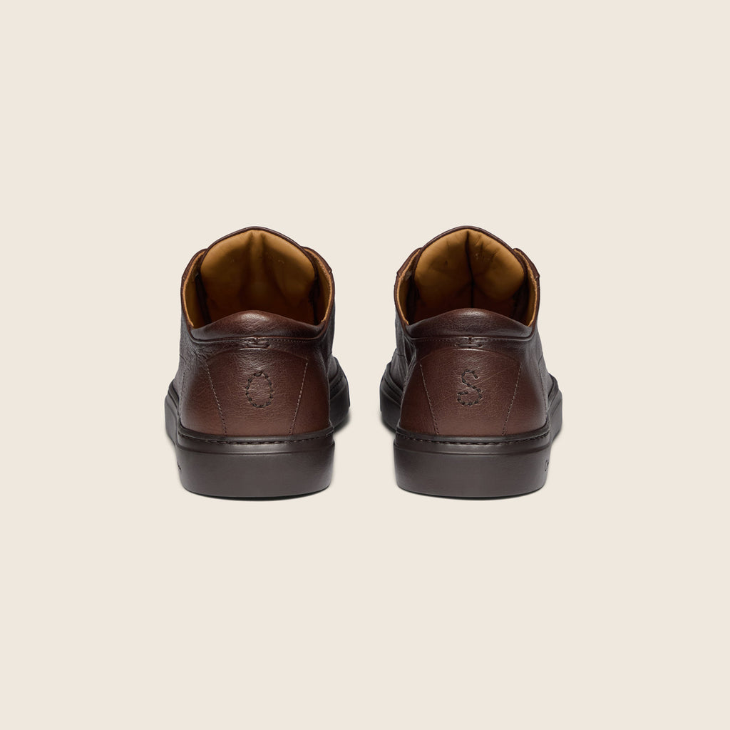 A pair of dark brown leather lace-up shoes shown from the back, featuring a textured leather heel counter, dark rubber soles, and a tan interior lining, set against a solid light beige background. Oliver Sweeney Shoes are available at Sakks Menswear in Southampton.