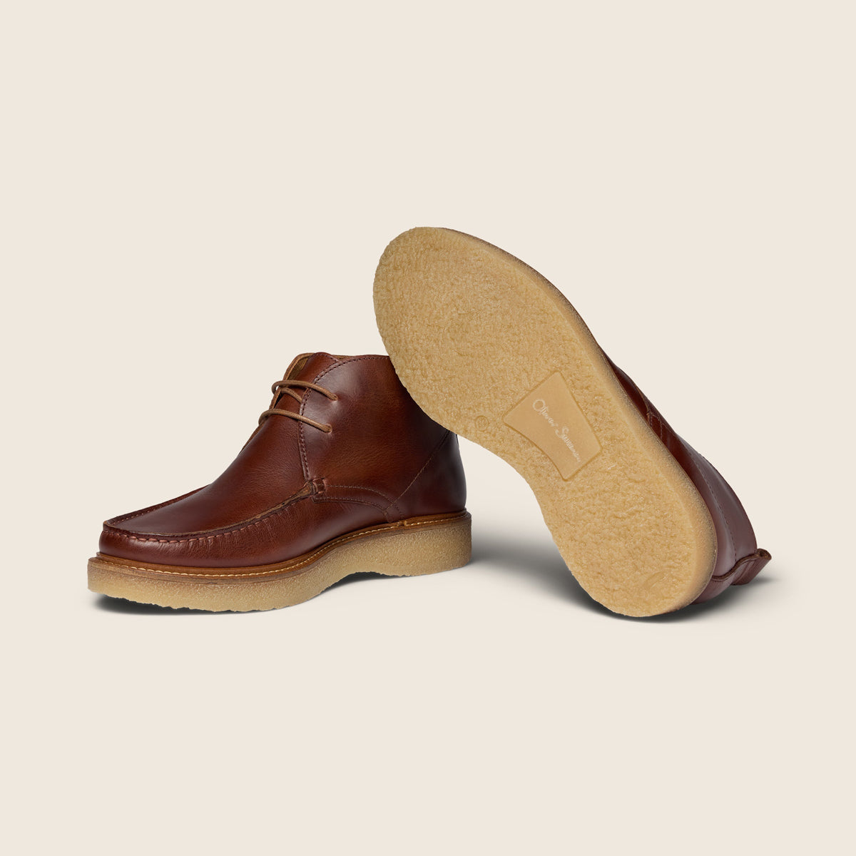 A pair of brown leather lace-up boots shown on a light beige background, with one boot standing upright to display the side and the other lying on its side to show the tan suede-like sole. Oliver Sweeney Moccasin Boots Available at Sakks Menswear in Southampton.