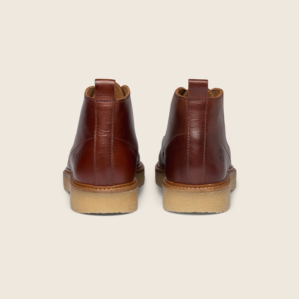 A pair of brown leather ankle boots shown from the back, featuring a tan rubber sole, set against a solid light beige background. Oliver Sweeney Moccasin Boots Available at Sakks Menswear in Southampton.