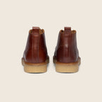 A pair of brown leather ankle boots shown from the back, featuring a tan rubber sole, set against a solid light beige background. Oliver Sweeney Moccasin Boots Available at Sakks Menswear in Southampton.
