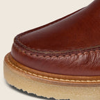 Close-up detail of a brown leather boot toe showing the stitched edge, leather grain, and a tan rubber sole against a light beige background. Oliver Sweeney Moccasin Boots Available at Sakks Menswear in Southampton.