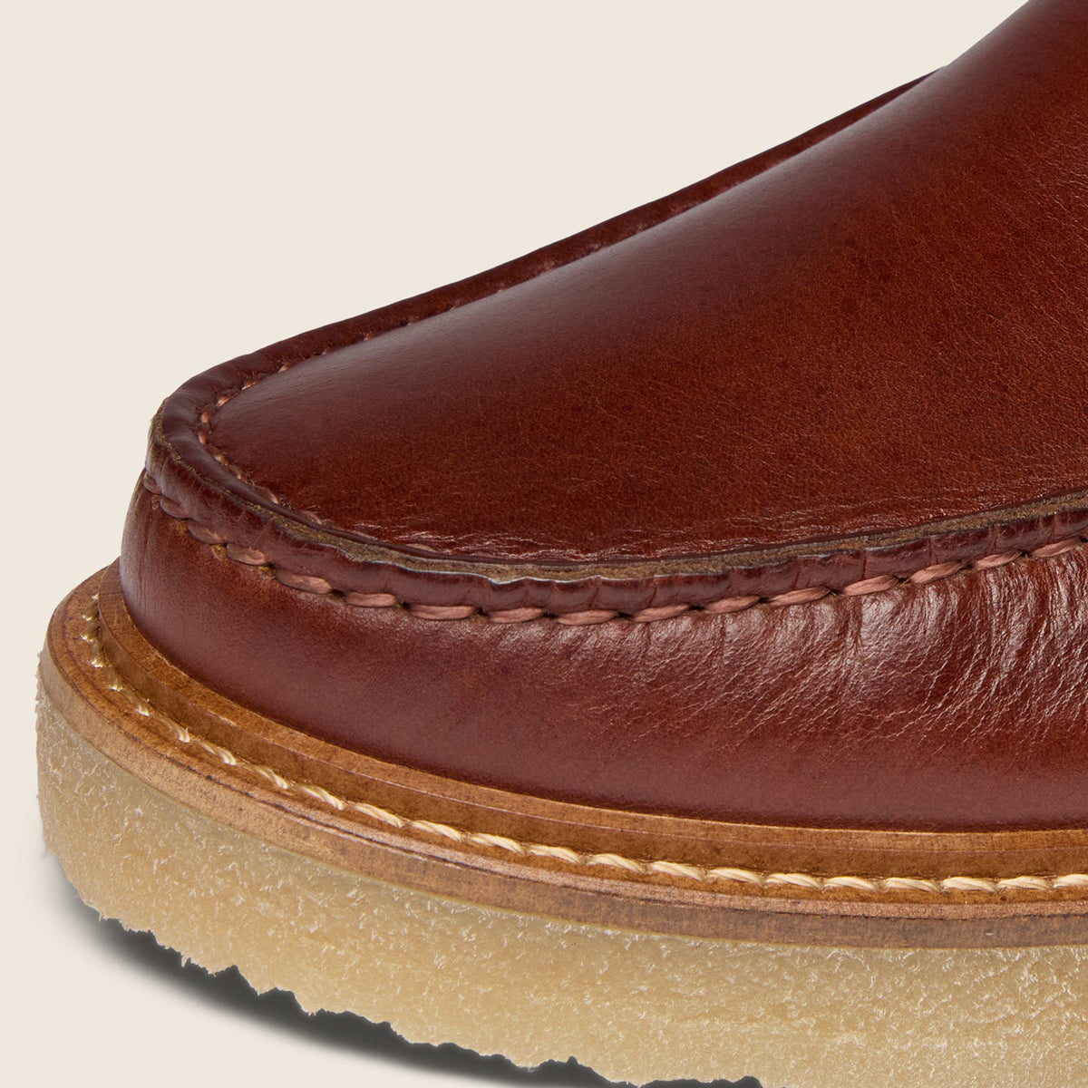 Close-up detail of a brown leather boot toe showing the stitched edge, leather grain, and a tan rubber sole against a light beige background. Oliver Sweeney Moccasin Boots Available at Sakks Menswear in Southampton.