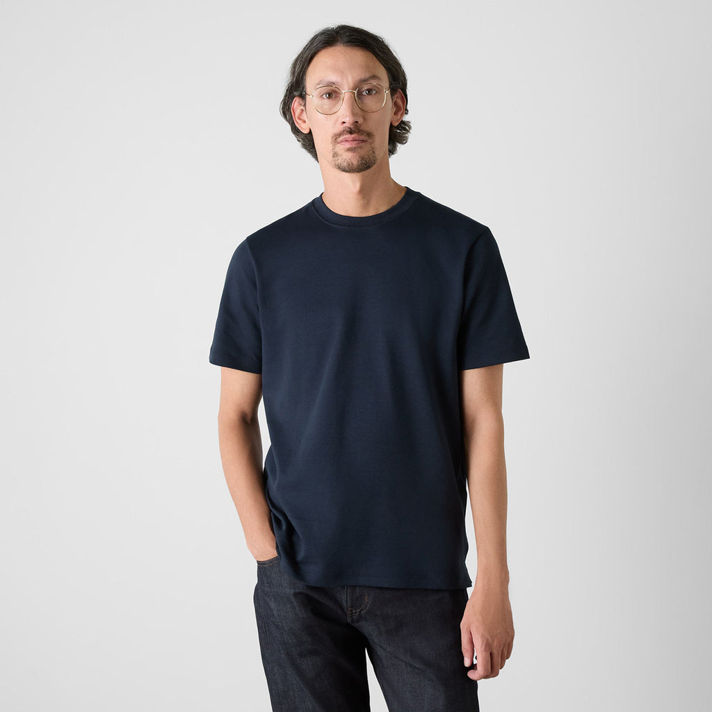 A man wearing a plain navy blue short-sleeve crewneck t-shirt and dark jeans, standing against a light beige studio background. Oliver Sweeney T-Shirts available at Sakks Menswear in Southampton.