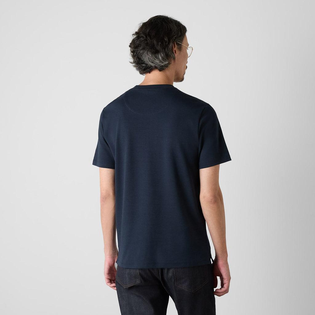 A man viewed from behind wearing a dark navy blue short-sleeve t-shirt and dark jeans, standing against a light gray studio background. Oliver Sweeney T-Shirts available at Sakks Menswear in Southampton.
