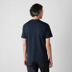 A man viewed from behind wearing a dark navy blue short-sleeve t-shirt and dark jeans, standing against a light gray studio background. Oliver Sweeney T-Shirts available at Sakks Menswear in Southampton.