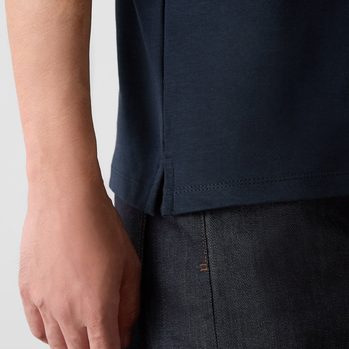 Close-up of a person wearing a dark navy blue short-sleeve t-shirt and dark charcoal gray pants, showing the side seam and hem detail of the shirt. Oliver Sweeney T-Shirts available at Sakks Menswear in Southampton.