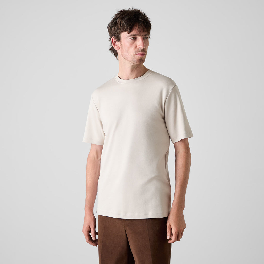 A man wearing a plain short-sleeve crewneck t-shirt in a light cream color, paired with brown trousers, standing against a light gray studio background. Oliver Sweeney T-Shirt available at Sakks Menswear in Southampton.