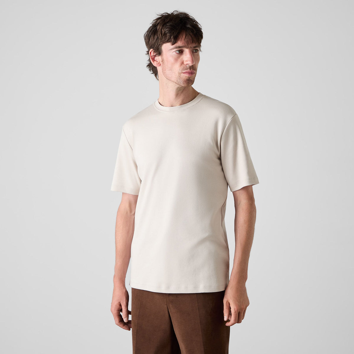 A man wearing a plain short-sleeve crewneck t-shirt in a light cream color, paired with brown trousers, standing against a light gray studio background. Oliver Sweeney T-Shirt available at Sakks Menswear in Southampton.
