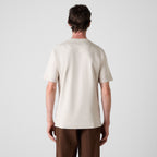 A man viewed from behind wearing a light cream/off-white short-sleeve crewneck t-shirt and brown trousers, set against a plain light gray studio background. Oliver Sweeney T-Shirt available at Sakks Menswear in Southampton.