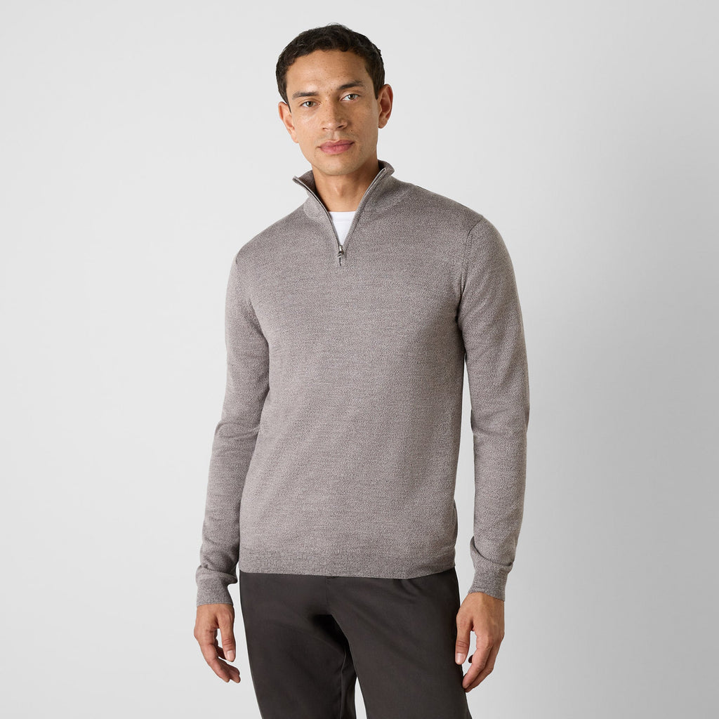 A man wearing a light gray heathered quarter-zip pullover sweater over a white undershirt and dark gray pants, standing against a light gray studio background. Oliver Sweeney Sweater available at Sakks Menswear in Southampton.