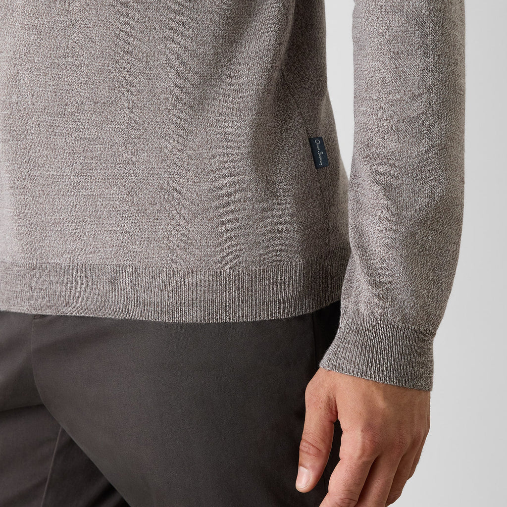 Close-up of a person wearing a heather gray knit sweater and dark trousers, showing the ribbed hem and cuff, with a small dark brand tab on the sleeve. Oliver Sweeney Sweater available at Sakks Menswear in Southampton.