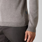 Close-up of a person wearing a heather gray knit sweater and dark trousers, showing the ribbed hem and cuff, with a small dark brand tab on the sleeve. Oliver Sweeney Sweater available at Sakks Menswear in Southampton.