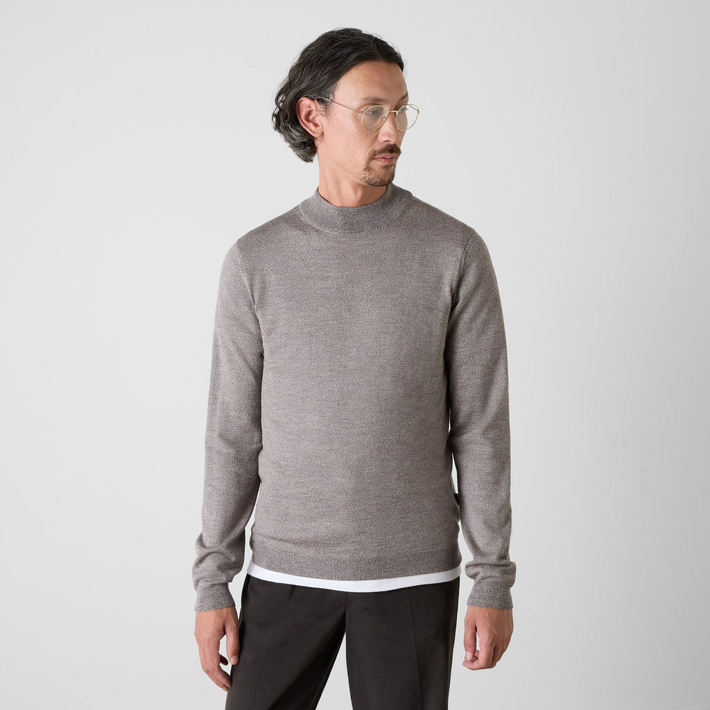 A man wearing a light gray mock neck knit sweater and dark trousers, standing against a light gray studio background. Oliver Sweeney Knitwear available at Sakks Menswear in Southampton.