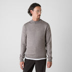 A man wearing a light gray mock neck knit sweater and dark trousers, standing against a light gray studio background. Oliver Sweeney Knitwear available at Sakks Menswear in Southampton.