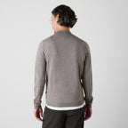A man viewed from the back wearing a heather gray mock-neck long-sleeve sweater over a white undershirt and dark trousers, set against a plain light gray studio background. Oliver Sweeney Knitwear available at Sakks Menswear in Southampton.