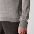 Close-up of a person wearing a heather gray knit sweater with dark trousers, showing the ribbed cuff and a small dark brand tag on the sleeve. Oliver Sweeney Knitwear available at Sakks Menswear in Southampton.