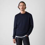 A man wearing a dark navy blue crewneck sweater, paired with dark jeans, standing against a plain light gray studio background. Oliver Sweeney Sweater available at Sakks Menswear in Southampton.