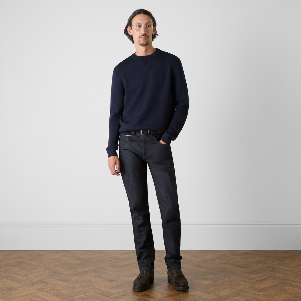 A man wearing a dark navy crewneck sweater, dark wash jeans, a black belt, and brown leather shoes, standing against a light gray wall on a wooden herringbone floor. Oliver Sweeney Sweater available at Sakks Menswear in Southampton.